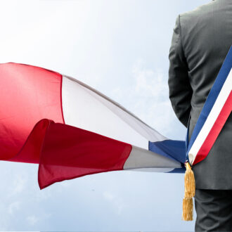 Un homme de so portant une écharpe d'élu aux couleurs de la France et un drapeau français.