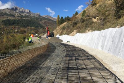 La scène se passe sur le chantier de construction d'une route. Au milieu de la photo des espèces de grillages sont posés sur le sol. En dessous on aperçoit du remblai gris. La route en travaux traverse la photo. Au second plan, une pelleteuse rouge est en train de répartir du remblai par dessus le grillage au sol. Au fond, un paysage de montagne à la végétation automnale. - Agrandir l'image 2 sur 3, fenêtre modale