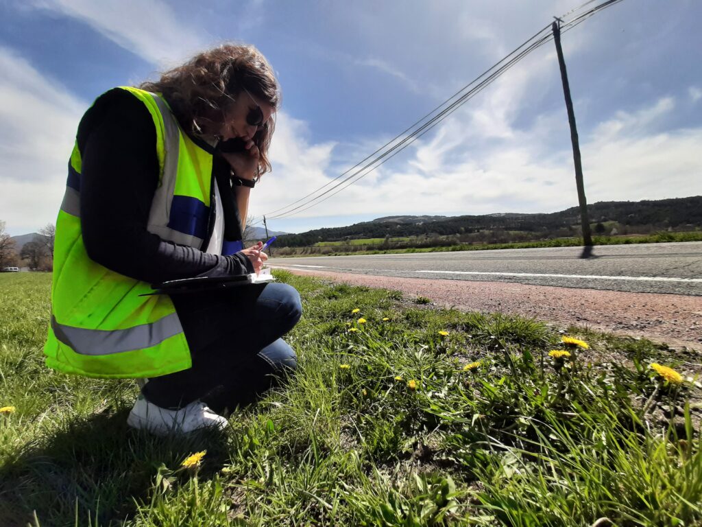 une agent en gilet sans manche jaune fluo par dessus un pull noir. Elle porte en jean foncé et des chaussures de sport blanche. Elle est accroupie téléphone à l'oreille et tablette sur les genoux en train de prendre des notes. Elle se situe sur la bas côté d'une route. - Agrandir l'image, fenêtre modale