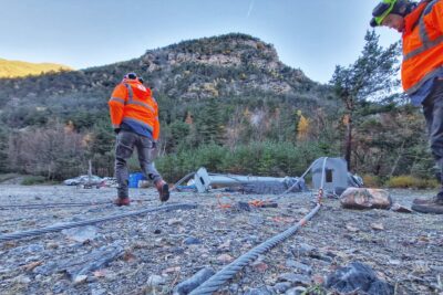 Un ouvrier en gilet orange en train de marcher à côté de poteaux métallique posés au sol en arrière plan des montagne - Agrandir l'image 21 sur 27, fenêtre modale