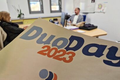Une affiche Duo Day apparait au premier plan. Au second plan, un agent en costume reçoit dans son bureau une participante au Duo Day. Ils sont en train de discuter - Agrandir l'image, fenêtre modale