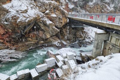 Dans un paysage, se distingue au milieu de la photo un torrent agité. Sur la berge sud, des blocs de béton en forme de Légo. En face une paroi rocheuse. Sur le côté droit de la photo, un pont traversant avec des garde-corps en métal gris. Il neige. Une pellicule blanche commence à se former. - Agrandir l'image 10 sur 12, fenêtre modale