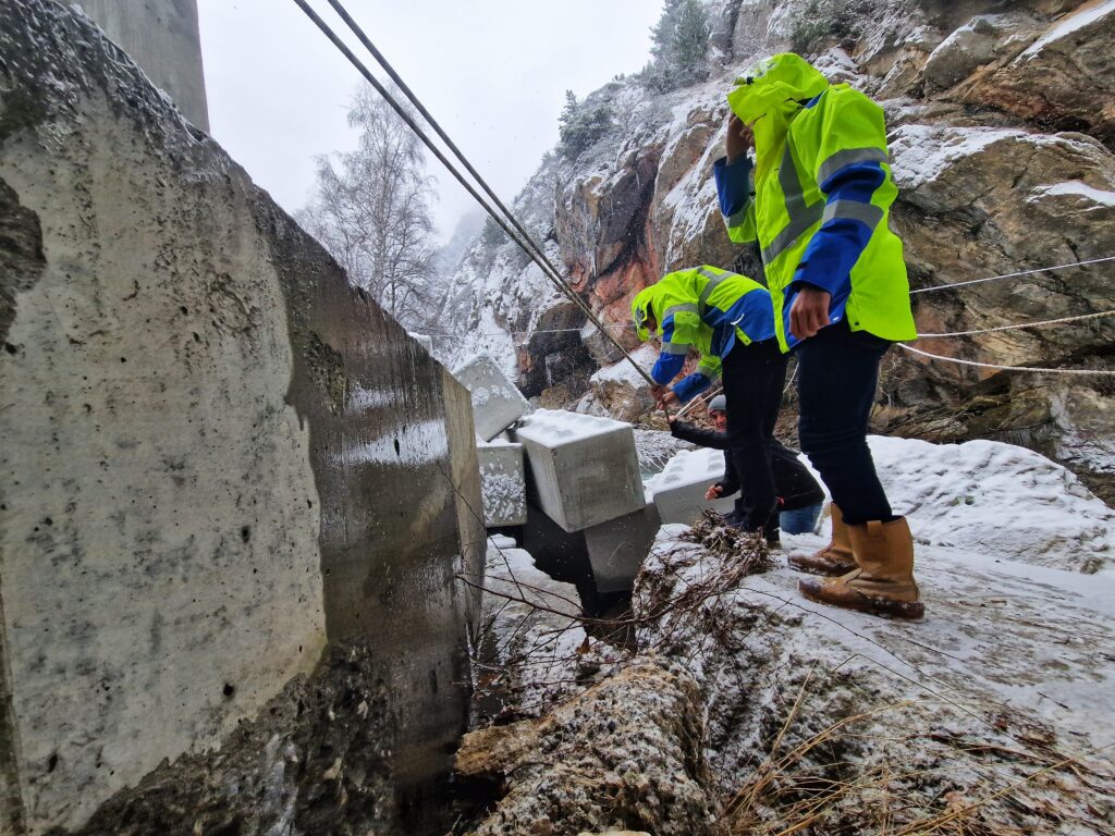 Deux agents du Département en habit de chantier jaune fluo inspectent la culée d'un pont. Au premier plan, se distingue un bloc de béton. Au second plan d'autres blocs de béton en forme de Légo s’enchevêtrent. En arrière plan une paroi de montagne rocheuse. - Agrandir l'image, fenêtre modale