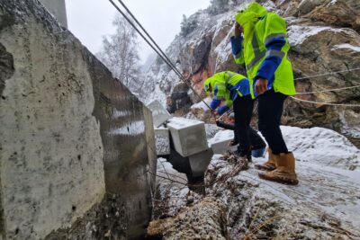 Deux agents du Département en habit de chantier jaune fluo inspectent la culée d'un pont. Au premier plan, se distingue un bloc de béton. Au second plan d'autres blocs de béton en forme de Légo s’enchevêtrent. En arrière plan une paroi de montagne rocheuse. - Agrandir l'image 9 sur 12, fenêtre modale