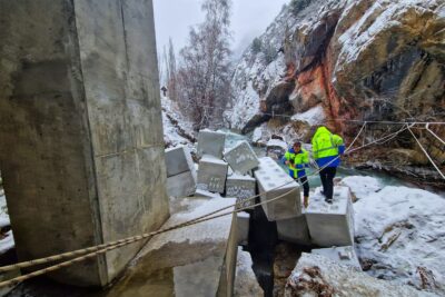 Au premier plan, le pilier rectangulaire en béton d'un pont. Juste derrière, des blocs de béton en forme de Légo. Sur celui le plus proche du torrent, deux homme en habit de chantier jaune fluo. Au milieu de la photo un torrent couleur bleu-vert. La berge d'en face se résume à une paroi rocheuse recouverte de rares conifères. Il neige, une pellicule blanche commence à se former. - Agrandir l'image 8 sur 12, fenêtre modale