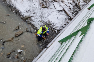 La photo est prise en plongée depuis le garde corps en métal vert d'un pont. En contre-bas, un homme habillée de jaune fluo est accroupi sur un ilot de terre au milieu du lit de la rivière et examine la partie inférieure du pont. Il neige à gros flocons. - Agrandir l'image 4 sur 4, fenêtre modale