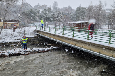 Sur un ilot au milieu du lit d'une rivière en furie, un agent du Département analyse la structure d'un pont chahuté par les eaux. Sur le pont une dale emmitouflée marche. À l'autre extrémité du pont un autre agent marche. Il neige à gros flocons. - Agrandir l'image 2 sur 4, fenêtre modale