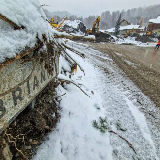 Au premier plan, un panneau en plein Risoul village sur lequel on peut lire Briançon se laisse apercevoir sous un amas de sédiments. La route est recouverte d'une couche de boue. En second plan, on distingue, sur la partie droite de la photo, un petit groupe d'hommes en habit de chantier jaune fluo. Sur le même plan, au milieu de la photo, des pelles mécaniques sont en train de défaire Risoul village de 30 000 m3 de sédiments. En arrière plan, on aperçoit la mairie. Il neige, une pellicule blanche est en train de se former.