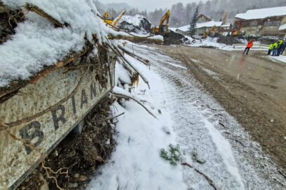 Au premier plan, un panneau en plein Risoul village sur lequel on peut lire Briançon se laisse apercevoir sous un amas de sédiments. La route est recouverte d'une couche de boue. En second plan, on distingue, sur la partie droite de la photo, un petit groupe d'hommes en habit de chantier jaune fluo. Sur le même plan, au milieu de la photo, des pelles mécaniques sont en train de défaire Risoul village de 30 000 m3 de sédiments. En arrière plan, on aperçoit la mairie. Il neige, une pellicule blanche est en train de se former. - Agrandir l'image, fenêtre modale