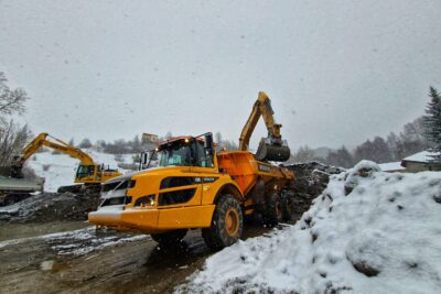 Une pelle mécanique remplit un imposant camion benne jaune muni de roues tout terrain. La pelle mécanique repose sur une impressionnante montagne de sédiments aux allures de gravats. Il neige, le paysage commence à blanchir. - Agrandir l'image 6 sur 12, fenêtre modale