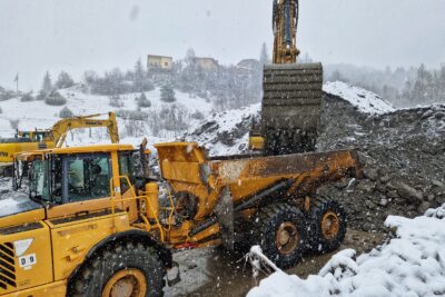 Une pelle mécanique remplit un imposant camion benne jaune muni de roues tout terrain. La pelle mécanique repose sur une impressionnante montagne de sédiments aux allures de gravats. Au fond, on aperçoit les chalets d'un hameau. Il neige, le paysage commence à blanchir. - Agrandir l'image 4 sur 12, fenêtre modale