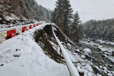 Photo de paysage sous la neige où l'on voit une route effondrée sur la moitié de la chaussée. Sur le coté droit de la route, un trou béant laisse apparaître les pieux de la glissière en bois suspendus dans le vide. Des protections de la route, rouge et blanc, protègent l'accès dangereux. En contre bas, le torrent du Réallon. En fond, la montagne et une forêt de conifères sous la neige. - Agrandir l'image 1 sur 12, fenêtre modale