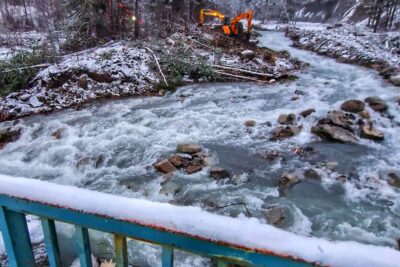Au premier plan, une partie des garde-corps d'un pont en métal couleur verdâtre. Au milieu de la photo, un torrent agité qui serpente. Sur la berge gauche, une pelle mécanique est en train de travailler sur font de paysage composé de conifères et de montagnes embrumées dans le fond. - Agrandir l'image 11 sur 12, fenêtre modale