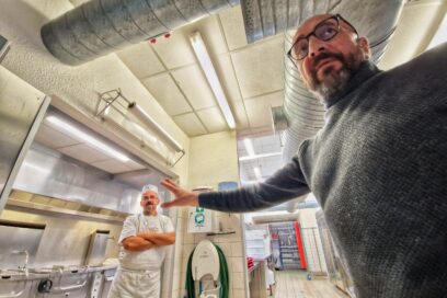 Au premier plan, un homme portant un pullover gris et des lunette au cerclage noir désigne de la main la pièce dans laquelle il se trouve. En l’occurrence une cuisine de collège au carrelage blanc. Au deuxième plan, un homme en habit et tablier blans qui porte un calot de cuisinier - Agrandir l'image, fenêtre modale
