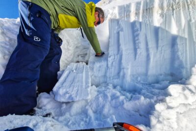 Un homme en tenue de ski est face à un petit mur de neige. Un bloc de taille moyenne s'est décroché de ce dernier. L'homme passe la main, penché en avant, sur la zone de fracture du bloc. Au premier plan on aperçoit le manche de sa pelle à neige. - Agrandir l'image 5 sur 13, fenêtre modale