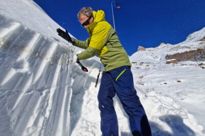 Face à un petit mur de neige, un homme en tenue de ski est en train de frapper sur le plat de sa pelle placée en haut d'une colonne découpée dans ce mur. Le ciel est bleu et lumineux - Agrandir l'image 7 sur 13, fenêtre modale