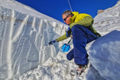 Accroupi devant un petit mur de neige, un homme en tenue de ski tend sa main en direction du mur. À coté de lui on distingue une pelle à neige. En arrière plan les montagnes. Le ciel est bleu et lumineux. - Agrandir l'image, fenêtre modale