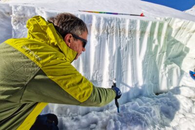 Un homme en tenue de ski, accroupi devant un petit mur de neige enfonce la lame de sa scie pliable dans la masse blanche, parallèlement au sol. Le ciel est bleu et lumineux. - Agrandir l'image 9 sur 13, fenêtre modale