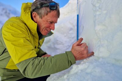 Dans un paysage blanc, un homme en tenue de ski se tient devant un petit mur de neige. Sur son côté, une sonde destinée à mesurer la hauteur de neige est planté. L'homme teste la dureté de la neige en plantant son doigt dans la partie supérieure du mur. - Agrandir l'image 11 sur 13, fenêtre modale