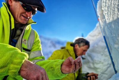Au premier plan, un homme béret sur la tête et imposantes lunettes de soleil noir sur le nez est habillé d'une polaire jaune fluo. Il est en train de tester la neige en faisant rouler au bout de ses doigts un petit échantillon prélevé sur place. En arrière plan, on distingue un homme en flou. Les deux hommes sont entourés de neige. Le temps est lumineux. Le ciel est bleu. - Agrandir l'image 12 sur 13, fenêtre modale