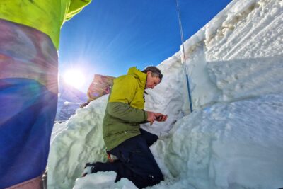 Au premier plan on distingue la jambe d'un homme habillé d'un pantalon bleu électrique est jeune fluo. Au deuxieme plan un homme est agenouillé dans la neige, face à un petit mur de neige. Il est en train d'entrer des données sur son smartphone. En face de lui, adossée au mur de neige, on aperçoit une sonde destinée à calculer la hauteur du mur. Le temps est lumineux. Le ciel est bleu. - Agrandir l'image 13 sur 13, fenêtre modale