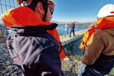 Au premier plan deux homme nous font dos. Ils portent un casque de chantier et des gilets de sauvetage. Au second plan, apparaît entre les deux hommes une passerelle en plastique gris clair flottante. Dessus deux autre personne, elles aussi portant un casque de chantier et un gilet de sauvetage marchent en direction de la marée de panneaux photovoltaïques qui se trouve en arrière plan. Au tout dernier plan, on distingue une ligne d'horizon montagneuse. Le ciel est bleu. - Agrandir l'image 1 sur 4, fenêtre modale