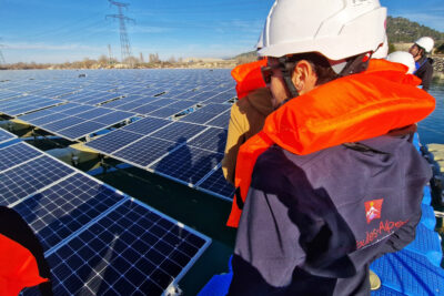Sur la droite de la photo, des personnes casque de chantier sur la tête et habillées d'un gilet de sauvetage sont en file indienne debout sur une espèce de ponton en plastique bleu électrique. Le ponton borde une marée de panneaux photovoltaïques flottants. En arrière plan, une ligne d'horizon montagneuse. - Agrandir l'image 3 sur 4, fenêtre modale