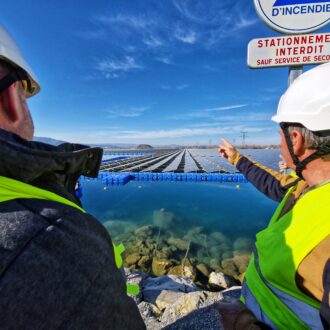 Deux hommes casque de chantier sur le tête et habillés d'un gilet jaune nous font dos au premier plan. Celui situé à droite désigne du doigt une marée de panneaux photovoltaïques qui flottent sur l'eau.
