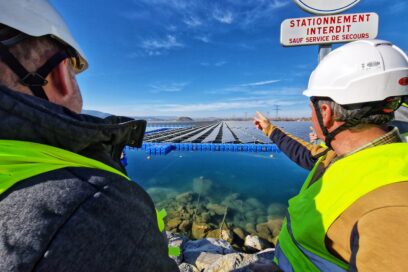 Deux hommes casque de chantier sur le tête et habillés d'un gilet jaune nous font dos au premier plan. Celui situé à droite désigne du doigt une marée de panneaux photovoltaïques qui flottent sur l'eau. - Agrandir l'image, fenêtre modale