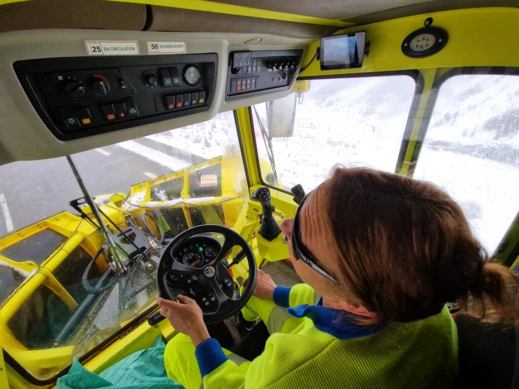 Une agent tout des jaune fluo vêtue, cheveux relevés dans un chignon et imposantes lunettes de soleil sur le nez. La photo est prise en plongée. L'agente est au volant d'un chasse neige dont la lame est en train de racler la chaussée revenue au noir. Sur les bas côté, de la neige. - Agrandir l'image, fenêtre modale