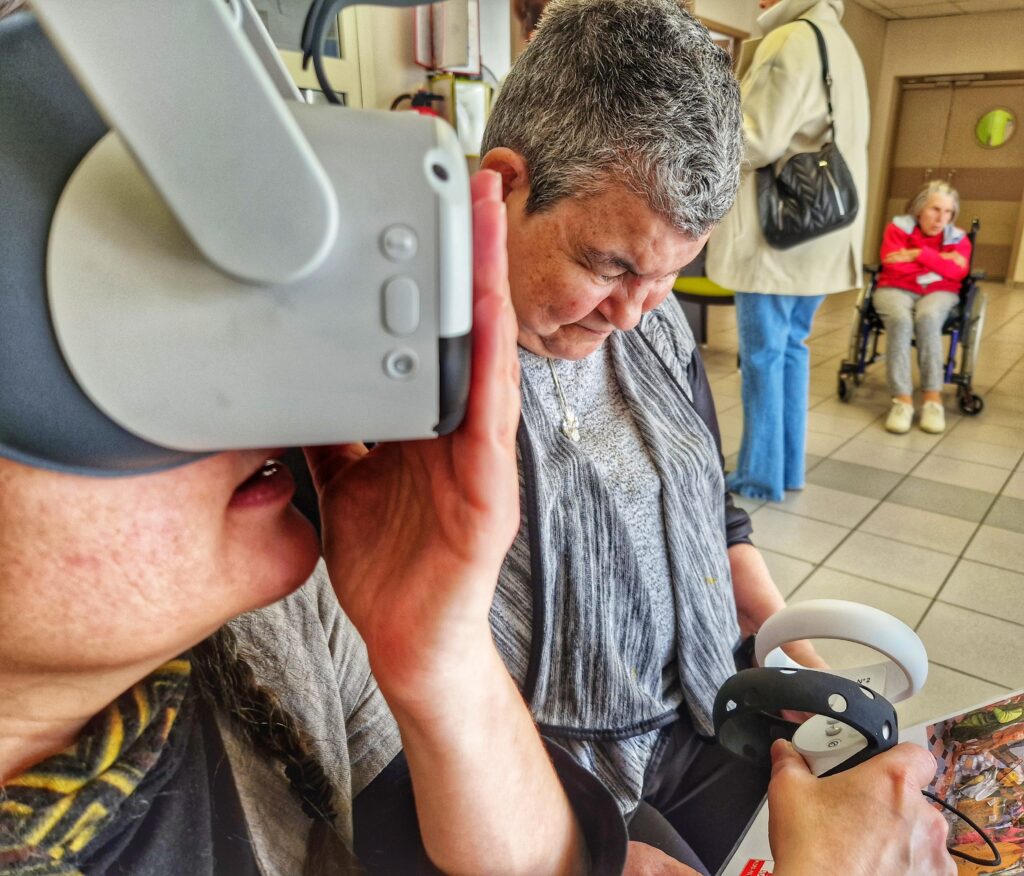 Au premier plan à gauche, gros plan sur le visage d'un personne portant un casque de réalité virtuelle. Juste à côté d'elle, le point est focus sur une dame d'un certain âge, les cheveux courts poivre et sel en train de lire une fiche posée sur ses genoux. Elle porte un pull gris chiné. En arrière plan on distingue une personne debout, portant un jean et un manteau beige et un sac à main noir sur son épaule droite. Encore un peu plus au fond, on distingue une dame âgée en fauteuil roulant. Elle porte une polaire rose et un bas de jogging gris. Ses bras sont croisés, elle regarde au loin/ La scène se passe dans le hall d'un Ehpad - Agrandir l'image, fenêtre modale