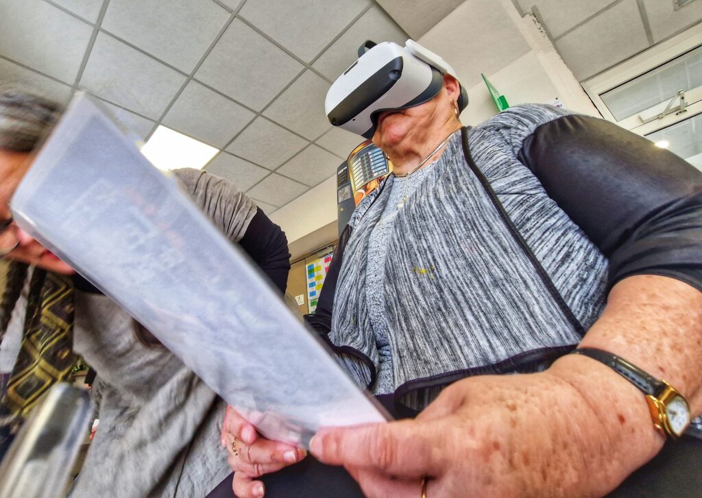 La photo est prise en contre-plongée. On y voit une dame d'un certain âge portant un pull gris chiné avec un casque de réalité virtuelle sur la tête. Elle tient également la fiche explicative du filme en court de visionnage. La scène se passe dans le hall d'un Ehpad. - Agrandir l'image, fenêtre modale
