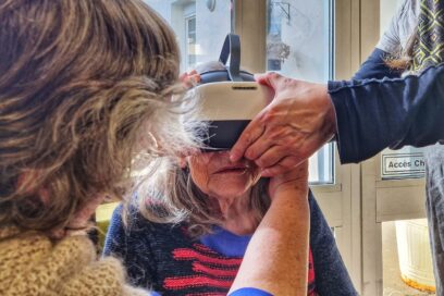 Au premier plan on voit une dame de dos; cheveux assez court mais faisant quelques bouclettes en train de mettre un casque de réalité virtuelle sur une dame d'un grand âge, qui occupe le centre de la photo. Une autre paires de mains est également présente pour aider à ajuster le casque. - Agrandir l'image, fenêtre modale