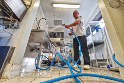 La scène se passe dans une cuisine de collectivité ou l'inox règne en maître. Au centre, un homme habillé en chef cuisinier tien un tuyau de couleur bleu dans ses main et arrose le sol de la cuisine fait de carrelage clair. - Agrandir l'image, fenêtre modale