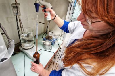 Une jeune fille rousse habillée d'une blouse blanche se tien devant un plan de travail accolé à un mur. Elle prélève dans un flacon un liquide à l'aide d'une pipette dont la partie qu'elle tient à la main à une forme de pistolet. Juste derrière le flacon en verre brun, un récipient est posé sur un petit promontoire. À l'intérieur, de l'eau tourbillonne. Afin d'en contrôler la température, une sonde en métal est plongé en son sein. - Agrandir l'image 3 sur 6, fenêtre modale