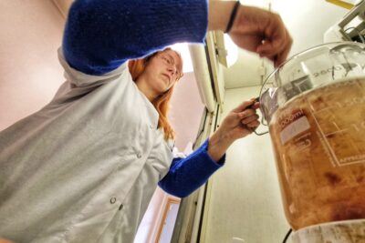 Une jeune femme rousse, cheveux longs, fines lunettes sur le nez est habillée d'une blouse blanche. En dessous, elle porte un pull bleu électrique dont les manches dépassent de la blouse. Elle est en train de verser une substance dont on ne voit pas le contenu dans un récipient en verre transparent rempli d'un liquide jaunâtre parsemé d'une mixture rouge (de la viande mixée). La photo est prise en contre plongée dans une ambiance de laboratoire. - Agrandir l'image 4 sur 6, fenêtre modale