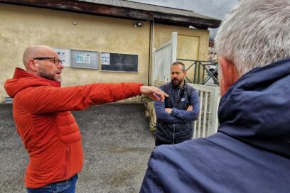 On distingue trois hommes dont un de dos au premier plan. Il porte une veste de pluie bleu marine, ses cheveux sont courts et blancs. Au second plan, une autre homme le crâne rasé et des lunettes à la monture noire visées sur le nez. Il porte une veste de montagne corail et un jean bleu foncé. Il tend le bars pour désigné le portail que l'on devine à deux pas du trio. Un peu plus vers le fond, le troisième homme, veste à manche courte bleu marine et chemise bleu plus claire. Il est brun, porte les cheveux courts. Une barbe fournie occupe le bas de son visage. Il croise les bras et porte une attention particulière à l'homme à la veste corail qui est en train de parler. Les scène se déroule dans l'enceinte d'un collège au niveau du portail d'entrée en métal blanc. Le temps est gris et pluvieux. Le bitume du sol est mouillé. - Agrandir l'image, fenêtre modale