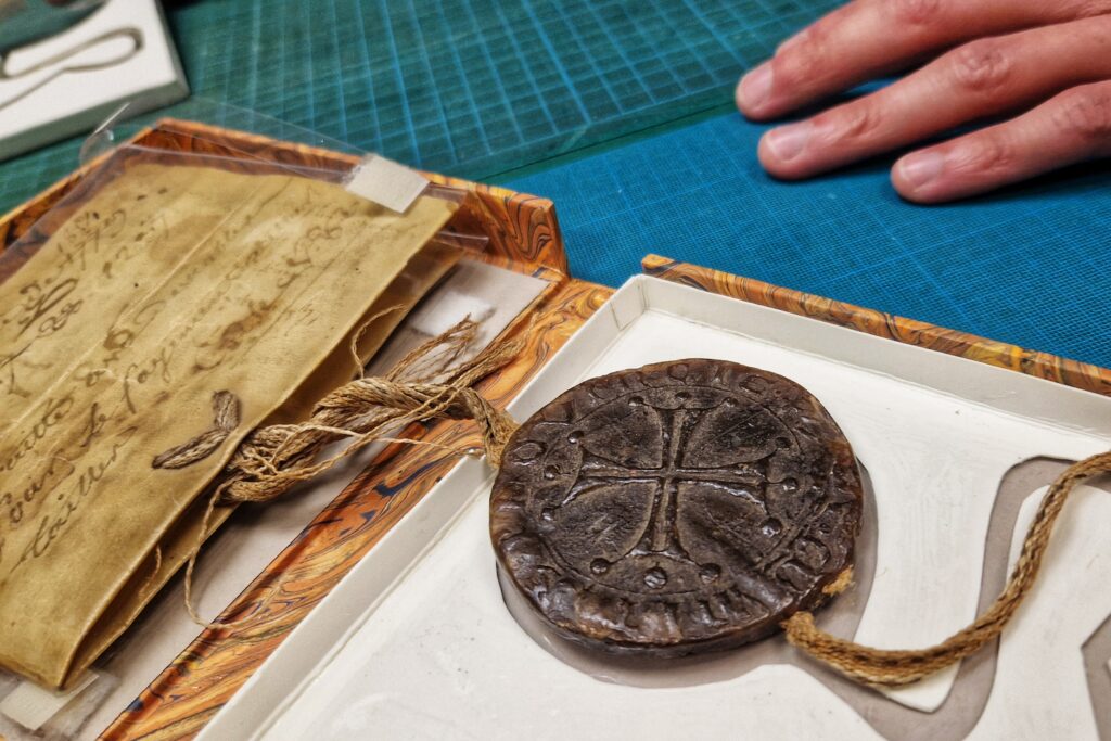 Dans une boite de conservation cartonnée un sceau en cire brune représente un croix occitane. Il est accroché par un filament à un parchemin en peau animal protégé dans une enveloppe plastique transparente. - Agrandir l'image, fenêtre modale