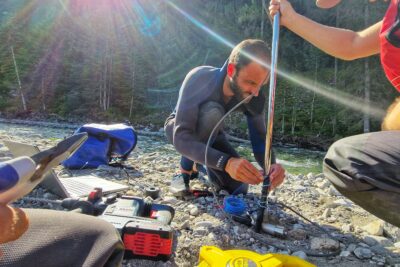 Au centre de la photo, un homme en combinaison de plongée de couleur sombre est en train de fixer une sonde (une espèce de tube noir) à un manche métallique. Il est accroupi. À coté de lui, sur la droite de la photo, une jeune fille elle-même accroupie, casquette sur la tête, lunettes de soleil sur le nez et gilet de sauvetage rouge, lui donne un coup de main. La scène se passe au bord du Drac,. Le temps est au soleil. - Agrandir l'image 9 sur 12, fenêtre modale