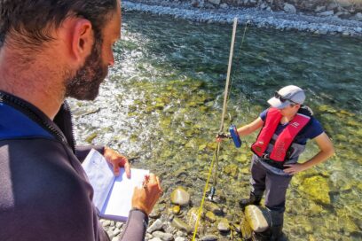 La scène se passe au bord du Drac,aus eaux bleu vert. Les pieds dans l'eau, une jeunes femme, habillée de cuissardes de couleur sombre, casquette sur la tête et gilet de sauvetage rouge. Elle tient un manche métallique équipé d'un boitier électronique dont elle est en train de lire des données. Sur la berge, une homme en combinaison de plongée tient un carnet sur lequel il est en train de noter ce que lui dicte la jeune fille. Le temps est ensoleillé - Agrandir l'image, fenêtre modale