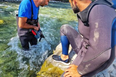 Sur la photo deux hommes. Celui de gauche est dans une rivière aux eaux vives bleu vert, le Drac. Il est en cuissardes, de l'eau jusqu'à mi-cuisses et tient dans sa main un perforateur dont une partie de la mèche est immergée dans l'eau. Il s'apprête à perforer un rocher, lui aussi plongé dans l'eau. Sur sa partie émergée, un autre homme en combinaison de plongée est assis. Il regarde l'homme à au perforateur. Le temps est ensoleillé. - Agrandir l'image 3 sur 5, fenêtre modale