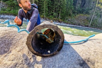 Au premier plan, dans un tube en PVC noir, un petit boitier rond et translucide renfermant des composants électroniques. Le tout est posé sur un rocher sur lequel s'appui un homme en combinaison de plongée. Au second plan, une rivière aux eaux vert bleu. Au fond, la berge et une forêt de résineux. Le temps est ensoleillé - Agrandir l'image 1 sur 5, fenêtre modale