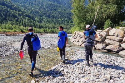 Dans le lit d'une rivière fait de méandres d'eaux bleu vert et des cailloux, trois personne marchent côté à côte nous faisant dos. Celle de droite, est un homme en combinaison de plongée, portant un cahier à la couverture rose rouge dans la mai droite et un sac à dos étanche couleur bleu sur l'épaule. Au milieu un autre hommes en cuissardes sombre porte un décamètre dans sa main droite et un sac à dos étanche bleu sur l'épale droite. À gauche, une jeune fille casquette sur la tête et en cuissardes sombre laisse reposer sur son épaule droite un instrument de mesure : une longue perche métallique surmonté d'un boitier électronique. - Agrandir l'image 2 sur 5, fenêtre modale