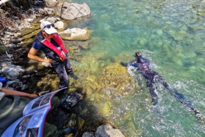 Dans les eaux bleu vert du Drac, un homme grenouille en combinaison néoprène noir nage. Sur la berge, une jeunes fille au cheveux courts, casquette sur la tête, habillée de cuissardes grises et d'un gilet de sauvetage rouge à les mains sur les hanche. Elle regarde la rivière. À côté d'elle, une autre jeune fille, en cuissardes vertes tient un porte document à la couverture rose. Sur les page visible, des schémas et des photos du lieu où a été prose la photo - Agrandir l'image 5 sur 5, fenêtre modale