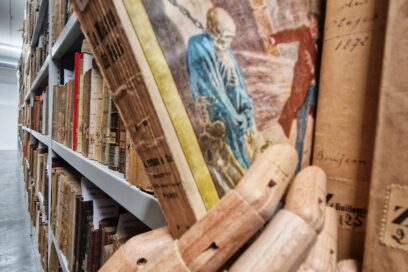 Au premier plan un main étrange en bois se saisit d'un livres dans le rayonnage de ce qui ressemble à une immense bibliothèque remplie de livre pour certains ancien. Le livre en question semble lui même assez ancien. Sur sa couverture, un squelette retenu par une chaine à son coup et un homme en redingote effrayé. - Agrandir l'image, fenêtre modale