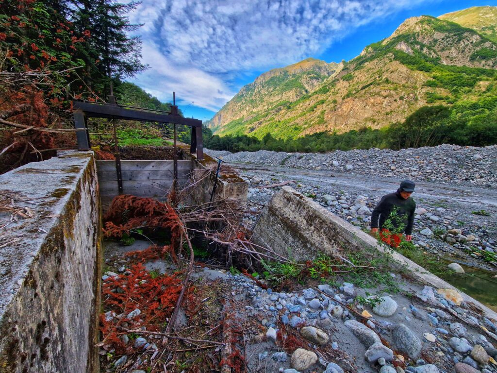 Au premier plan, la vanne d'un canal en béton complètement détruite et remplie de pierres, de sable, et de branchage. Juste à côté dans le large lit d'un rivière asséché, un homme en train de marcher. Il porte une veste marron gris, une casquette d'hiver que lui recouvre également les oreilles et un pantalon orange. Le lit de la rivière est caillouteux. Au loin, des montagne parsemé de vert. Le ciel est bleu et légèrement voilé de fins nuages. - Agrandir l'image, fenêtre modale