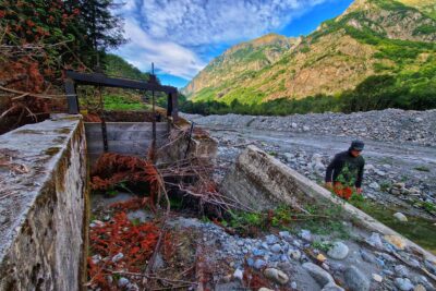 Au premier plan, la vanne d'un canal en béton complètement détruite et remplie de pierres, de sable, et de branchage. Juste à côté dans le large lit d'un rivière asséché, un homme en train de marcher. Il porte une veste marron gris, une casquette d'hiver que lui recouvre également les oreilles et un pantalon orange. Le lit de la rivière est caillouteux. Au loin, des montagne parsemé de vert. Le ciel est bleu et légèrement voilé de fins nuages. - Agrandir l'image 11 sur 12, fenêtre modale