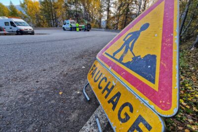 Au premier plan sur la parti droite de la photo un panneau triangulaire jaune travaux sous lequel on peut lire "Fauchage". Panneau situé sur le bord d'une route qui traverse la photo. En arrière plan une camionnette blanche munie qui tracte un broyeur de brache couleur verte. Sur son côté, des agents du Département habit de travail jaune fluo et bleu électrique. - Agrandir l'image 1 sur 7, fenêtre modale