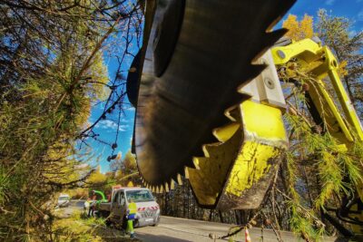 La photos est prise en contre plongée. Au premier plan, de très près une disque de métal au dents acérées. On devine qu'il est la partie final d'un bas mécanique articulé. Quel branchage sont prise dans le bras. Le disque est tout proche, de façon parallèle à des rangé d'arbres qui se trouve sur la bas côté d'une route. Les arbres sont des mélèzes. En arrière plan, une camionnette de chantier blanche devant laquelle se trouve, debout, un homme en vêtement de travail jaune fluo et bleu électrique. La camionnette tracte un broyeur couleur verte. Un peu plus en arrière, on distingue deux autres hommes en tenue de travail semblent porter un casque de chantier rouge. La photo est traversée en diagonale par une route. Le ciel est bleu parsemé de nuages. - Agrandir l'image 12 sur 12, fenêtre modale