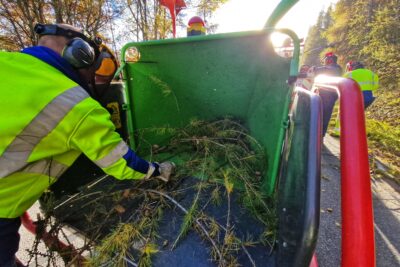 Sur partie droite de la photo un homme en vêtement de travail jeune porte un casque antibruit et une visière de protection comme grillagée. Il est train de pousser au fond d'un broyeur de branches couleur vert et rouge, des banchage de mélèzes. Un peu plus au fond, sur la partie droite de la photo on distingue deux hommes de dos également en vêtement de travail. Le temps est ensoleillé. - Agrandir l'image 7 sur 7, fenêtre modale
